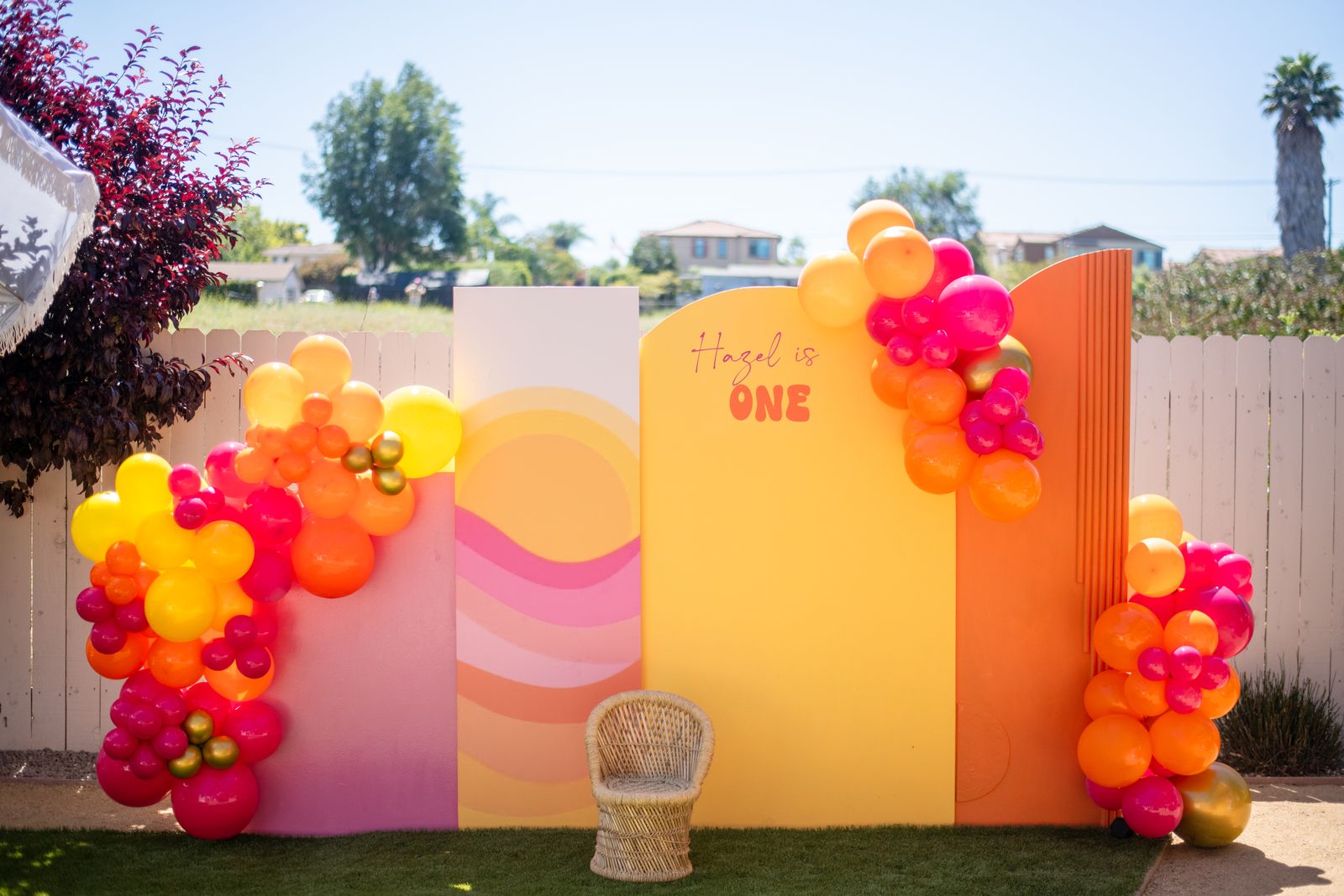 Orange and pink first birthday backdrop with balloon garland and a wicker chair